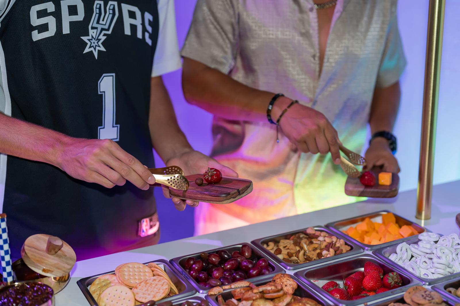 Two guests serving themselves strawberries and cheddar with gold tongs